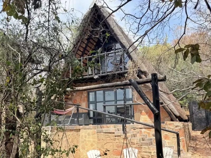 A rustic A-frame house featuring a thatched roof, stone exterior cladding, and a private balcony overlooking a natural garden setting.