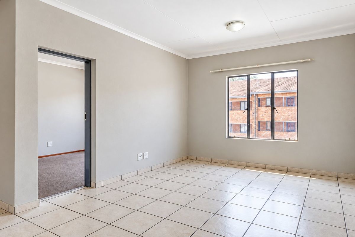 A bright, empty living area featuring neutral tiled flooring, clean walls, and a large window providing natural light and a view of the complex.