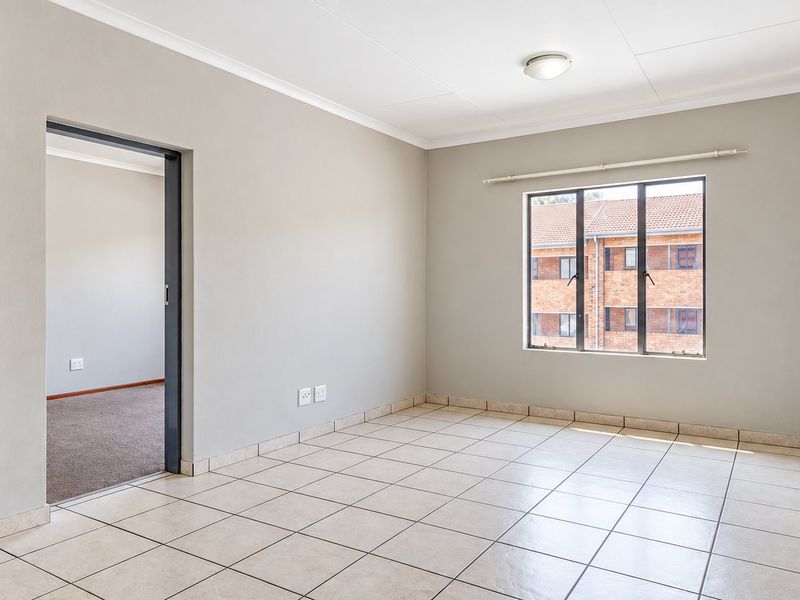 A bright, empty living area featuring neutral tiled flooring, clean walls, and a large window providing natural light and a view of the complex.