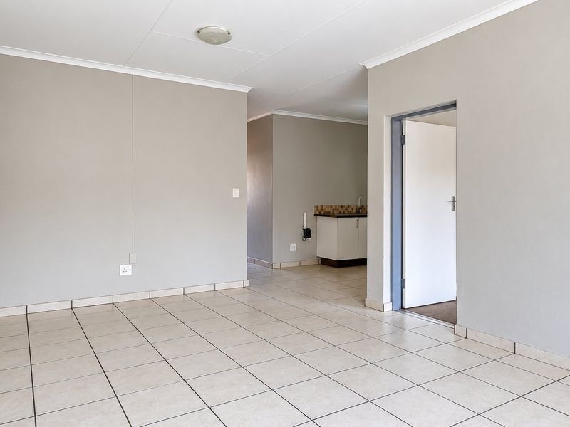 An empty open-plan living area featuring neutral tiled flooring, light grey walls, and a view into a compact kitchen space with built-in cabinetry.