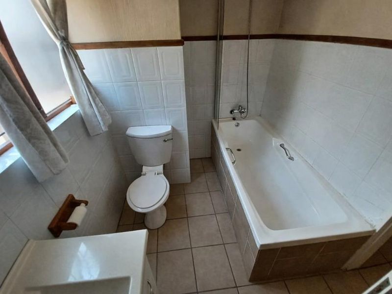 A functional bathroom featuring a bathtub with a shower mixer, glass screen, and toilet, finished with neutral wall and floor tiling.