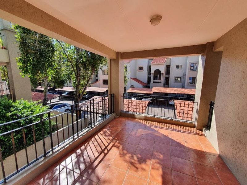 A spacious covered balcony featuring tiled flooring and wrought iron railings, offering an elevated view of the apartment complex grounds and parking area.