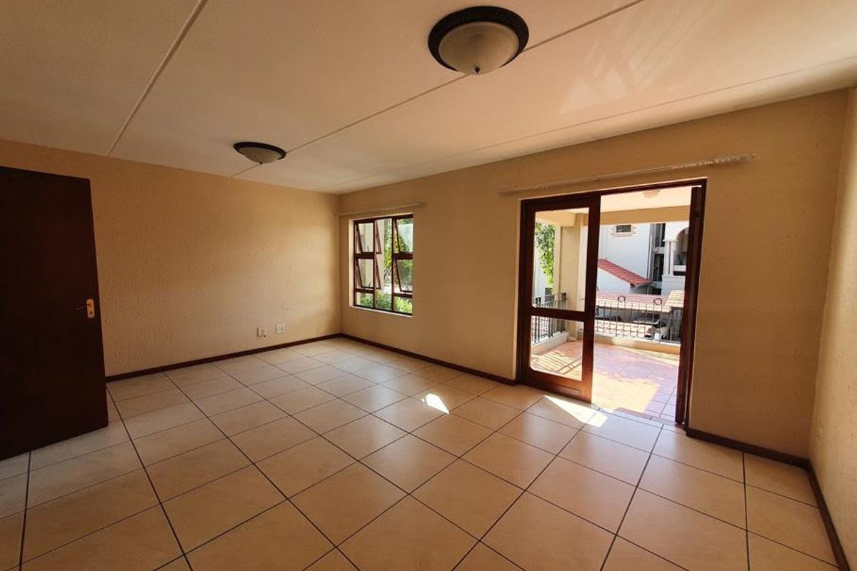 An empty, sunlit living room featuring neutral tiled flooring, flush mount lighting, and direct access to an outdoor balcony through a glass panel door.