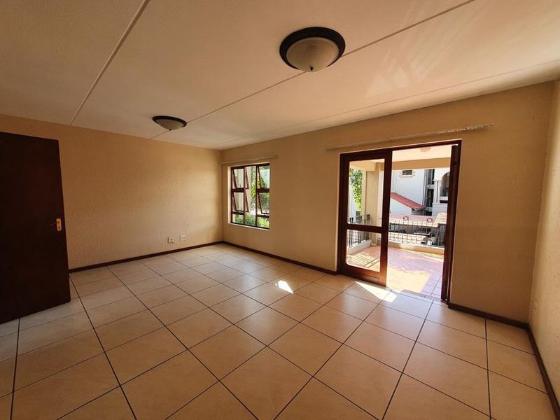 An empty, sunlit living room featuring neutral tiled flooring, flush mount lighting, and direct access to an outdoor balcony through a glass panel door.