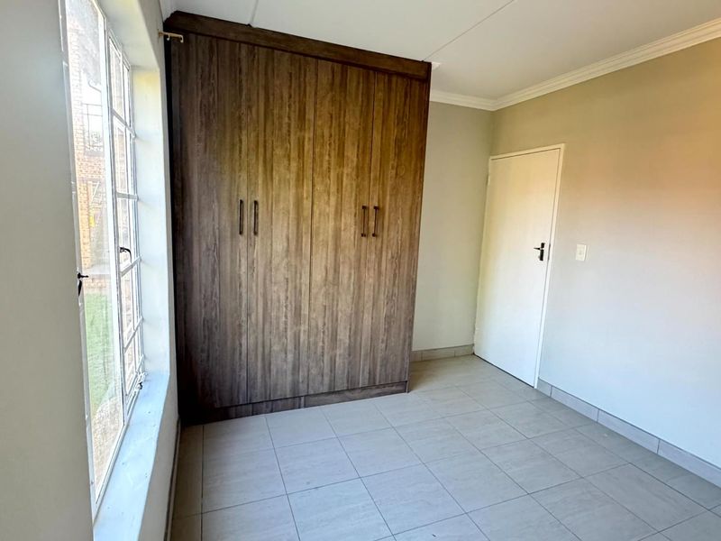 A bright, empty bedroom featuring neutral tiled flooring, built-in wooden cupboards, and a large window providing natural light.