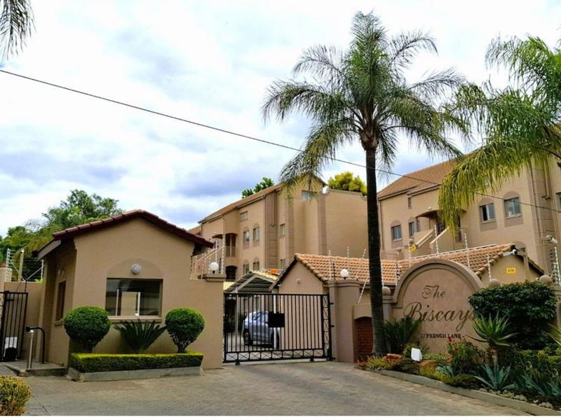 Front entrance view of The Biscays apartment complex, featuring a secure gated access point, perimeter wall with electric fencing, and landscaped garden beds.