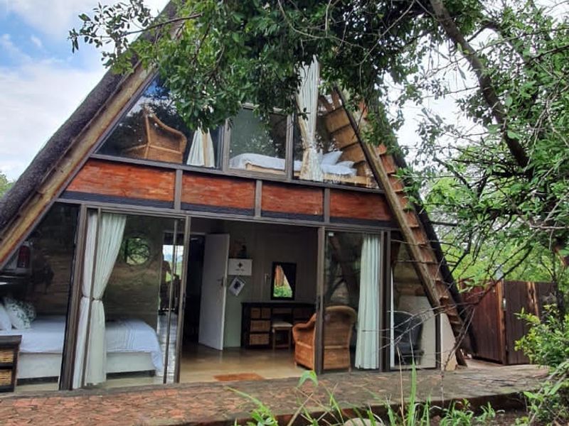 A unique A-frame house featuring a thatched roof, timber construction, and large glass sliding doors opening onto a brick-paved patio area.