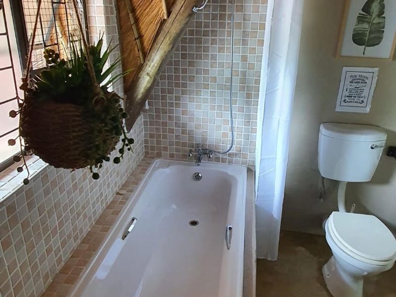 A rustic bathroom featuring a built-in bathtub, shower fixture, and toilet, set under a unique thatched roof with exposed timber beams.