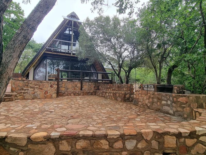 A rustic A-frame house featuring a large stone-paved patio with a built-in braai area, set within a natural, wooded environment.
