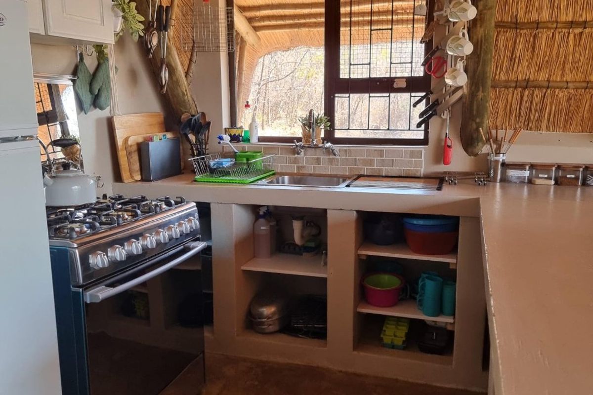 Rustic kitchen featuring a thatched roof, exposed timber beams, built-in concrete cabinetry, gas stove, and a stainless steel sink with garden views.