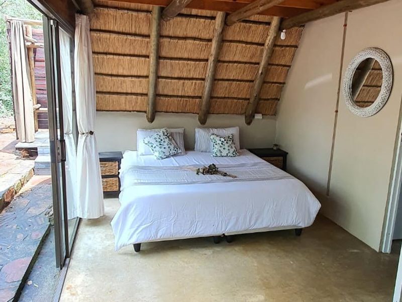 A rustic bedroom featuring exposed timber beams, a thatched roof ceiling, polished concrete floors, and large sliding glass doors opening to the outdoors.