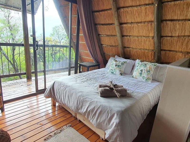 A rustic bedroom featuring exposed thatch roofing, timber flooring, and large sliding glass doors that open onto a private balcony with scenic views.