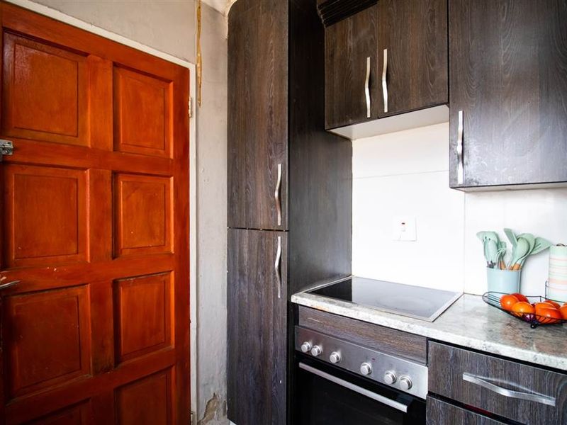 Compact kitchen area featuring dark wood-look cabinetry, integrated electric hob and oven, and stone-effect countertops, requiring some cosmetic wall repairs.