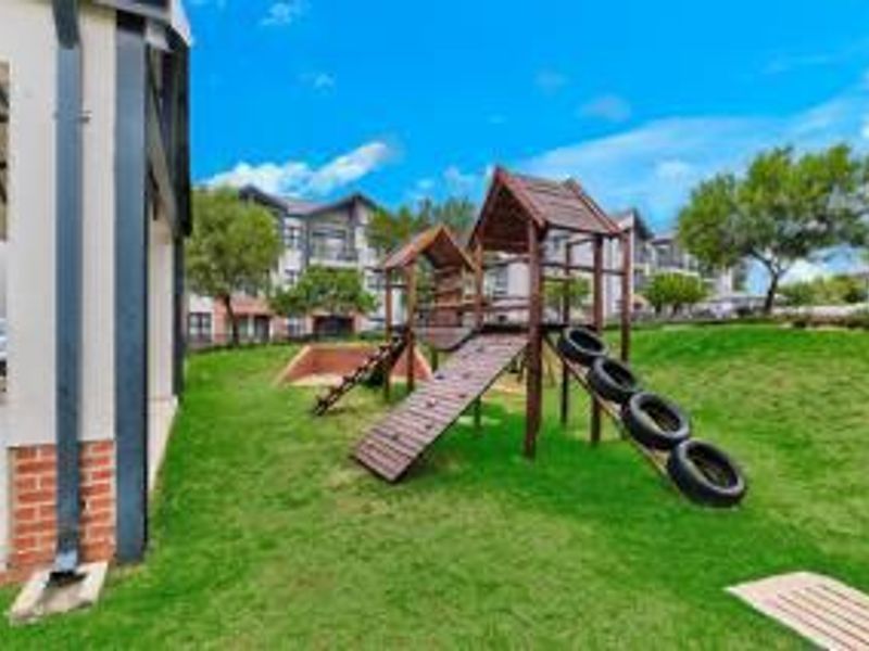 A communal outdoor area featuring a wooden playground structure set within a well-maintained lawn, adjacent to the apartment building exterior.