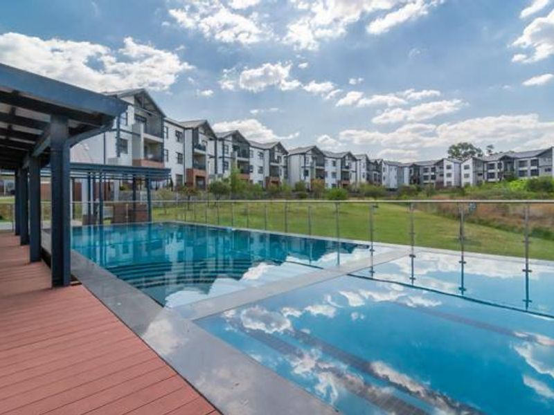 A modern swimming pool area featuring a sleek deck, glass balustrades, and a covered patio overlooking the apartment complex grounds.