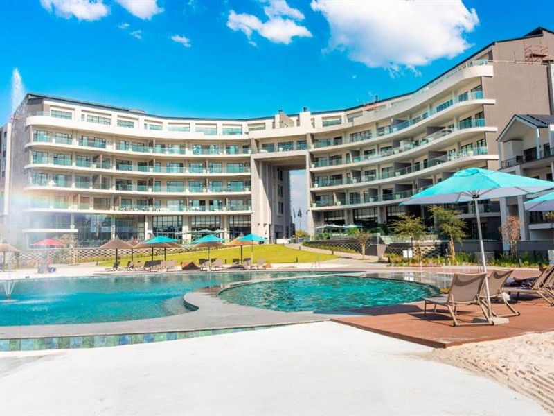A modern apartment complex featuring a large swimming pool, water fountain, landscaped grounds, and private balconies for residents.