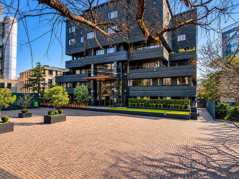 Modern apartment building exterior featuring a dark brick facade, paved entrance courtyard, glass canopy, and manicured landscaping.