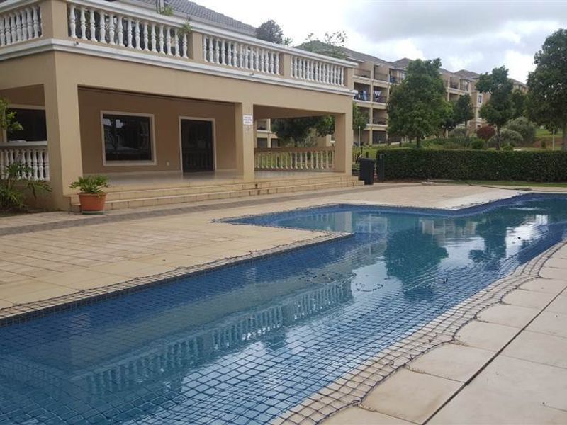 A communal swimming pool area featuring a safety net, tiled decking, and a covered patio with balustrades in a residential apartment complex.