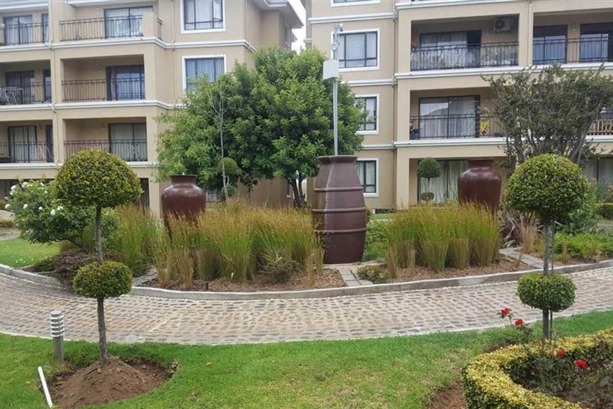 A well-maintained communal garden area featuring landscaped greenery, decorative urns, and cobblestone pathways, set against the backdrop of a multi-story apartment complex.