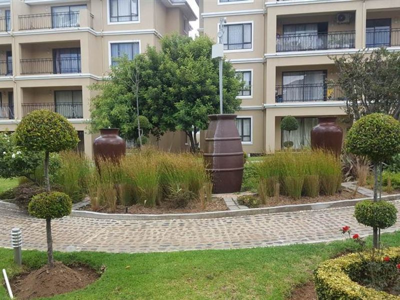 A well-maintained communal garden area featuring landscaped greenery, decorative urns, and cobblestone pathways, set against the backdrop of a multi-story apartment complex.