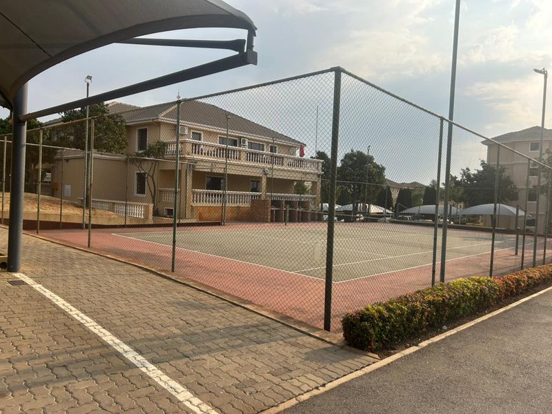 A well-maintained outdoor tennis court facility within the apartment complex, featuring perimeter fencing, paved access paths, and integrated floodlighting.