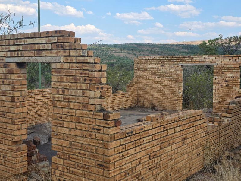 An unfinished brick structure on a vacant residential plot, featuring exposed masonry walls and a concrete slab foundation set against a natural landscape.