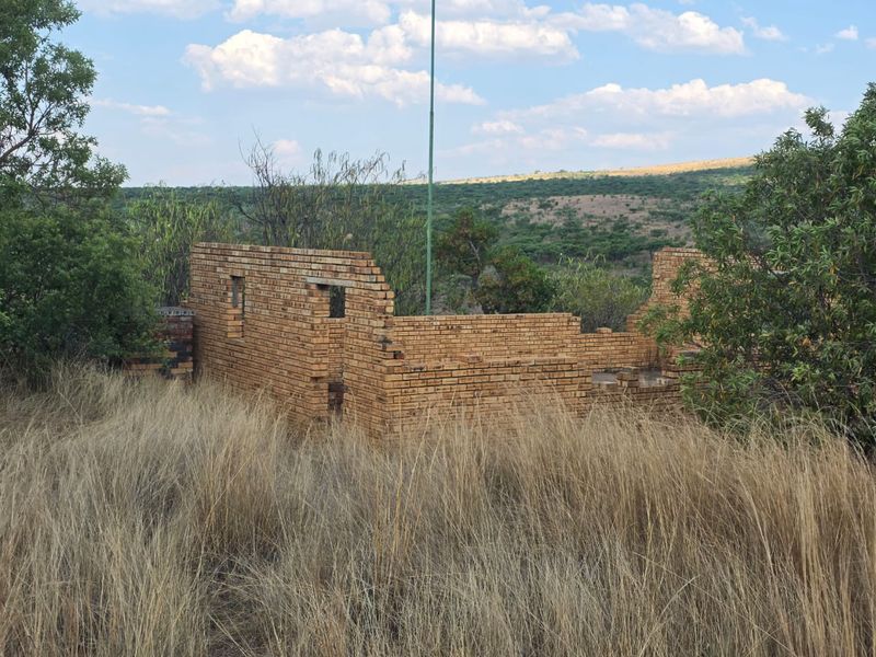 An incomplete brick structure situated on a vacant residential plot, surrounded by natural vegetation and open landscape.