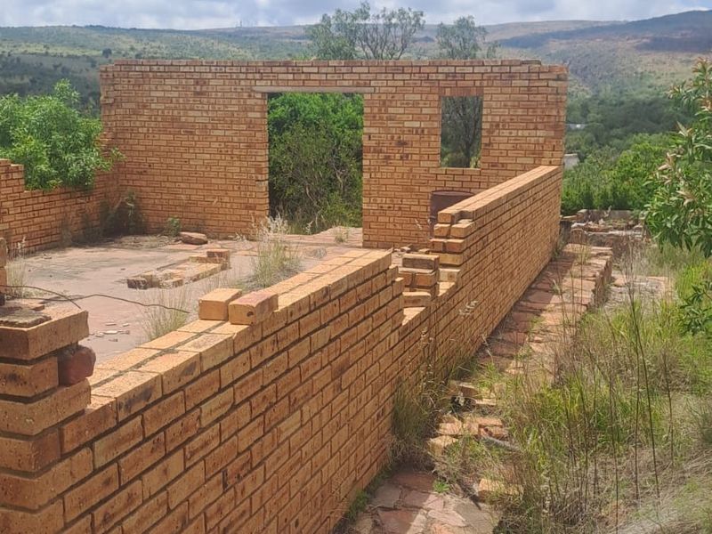 An unfinished brick structure on a residential plot, featuring exposed masonry walls and a concrete slab foundation set within a natural landscape.