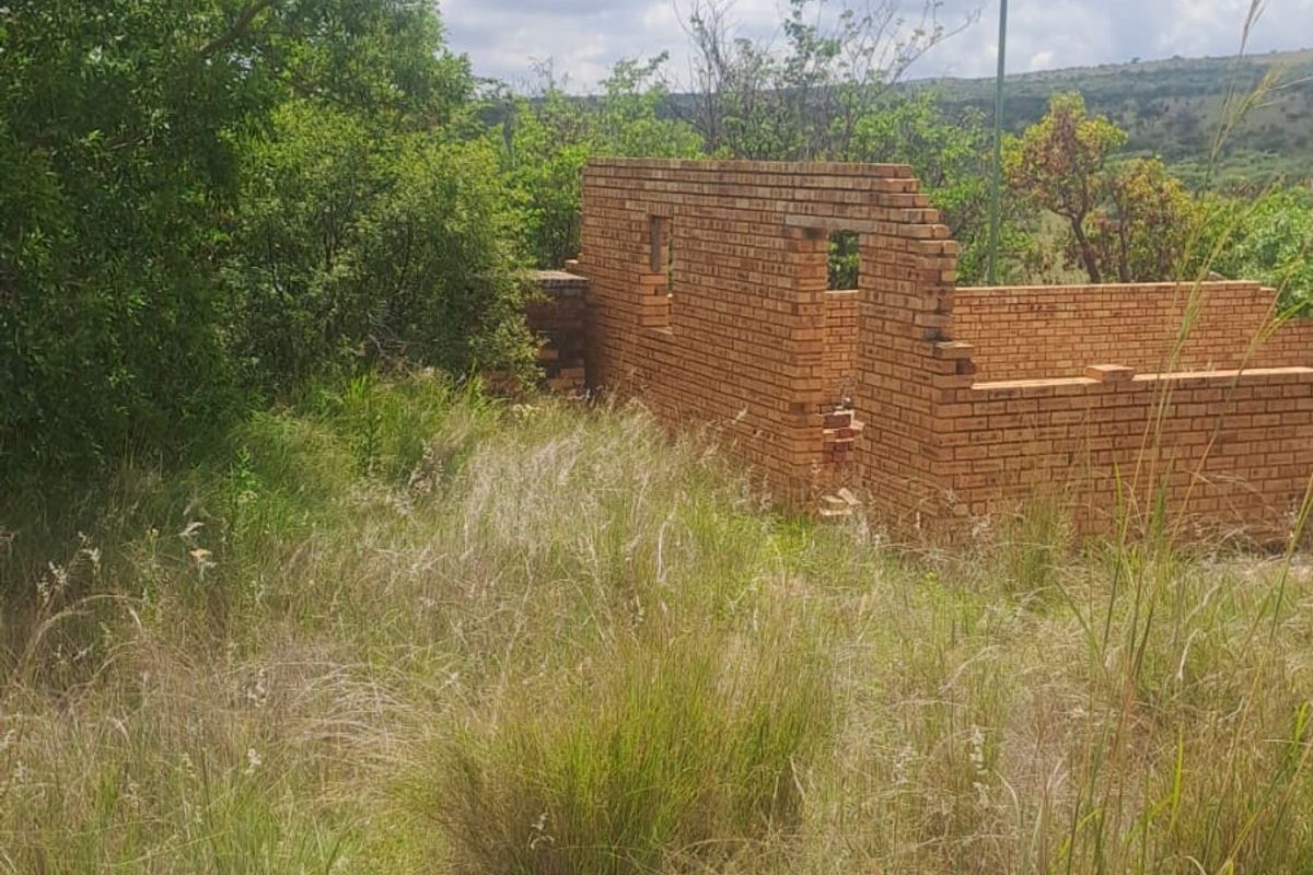 A view of an incomplete brick structure situated on a vacant residential plot surrounded by overgrown natural vegetation.