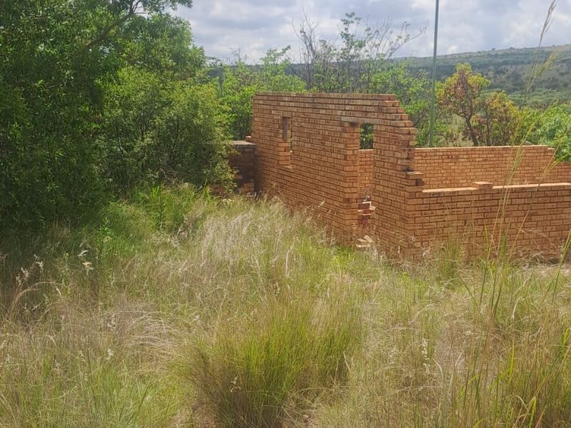 A view of an incomplete brick structure situated on a vacant residential plot surrounded by overgrown natural vegetation.