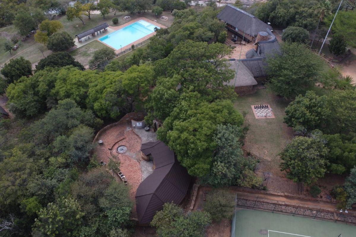 Aerial view of a spacious residential property featuring a swimming pool, tennis court, thatched roof buildings, and extensive landscaped grounds.