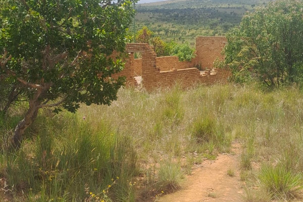 A view of an incomplete brick structure situated on a vacant residential plot surrounded by natural vegetation and hilly landscape.