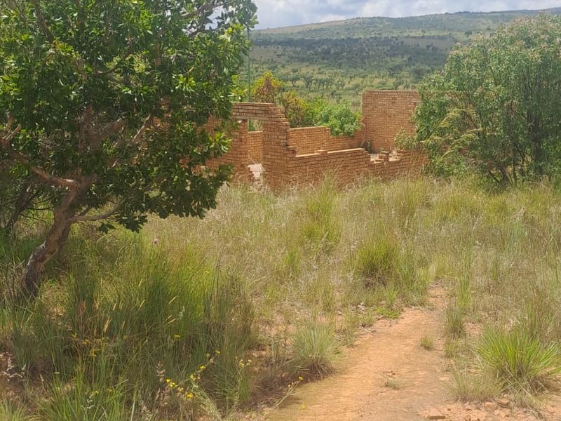 A view of an incomplete brick structure situated on a vacant residential plot surrounded by natural vegetation and hilly landscape.