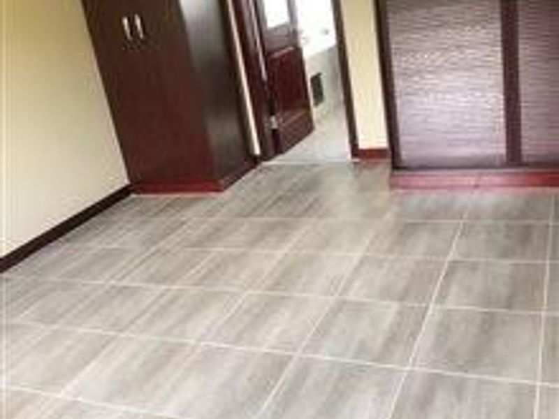 A clean, empty bedroom featuring modern wood-look tiled flooring and dark wood built-in cupboards, with an adjacent doorway leading to another room.