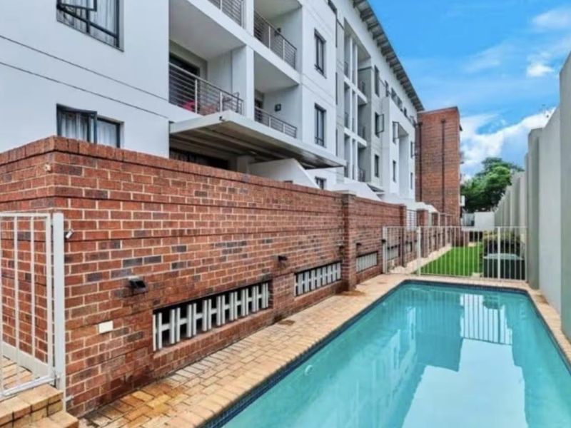 A well-maintained communal swimming pool area featuring a brick boundary wall, paved surround, and views of the apartment building balconies.