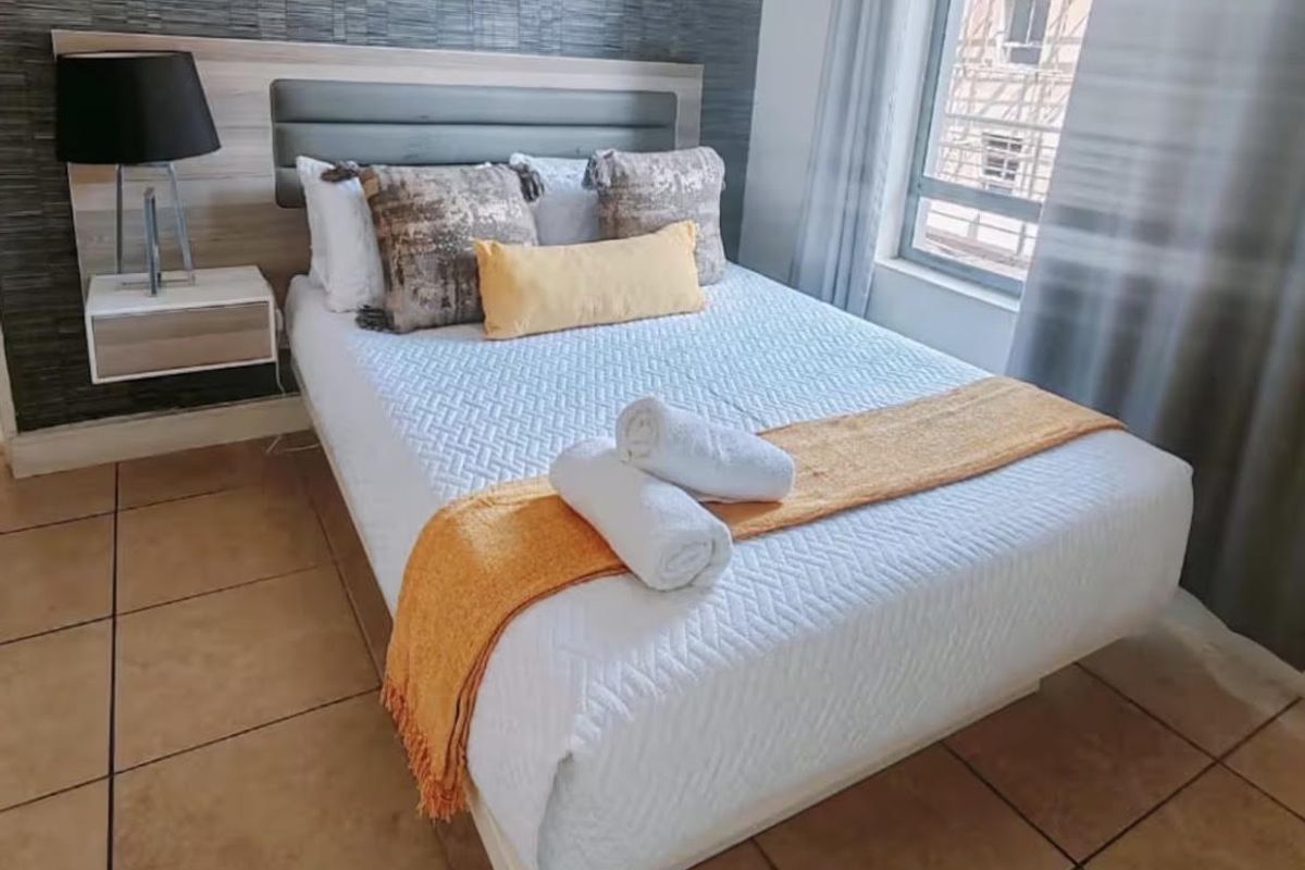 A well-lit bedroom featuring tiled flooring, a modern built-in headboard with a floating bedside table, and a large window providing natural light.