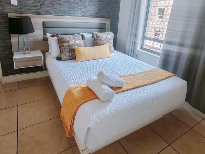 A well-lit bedroom featuring tiled flooring, a modern built-in headboard with a floating bedside table, and a large window providing natural light.