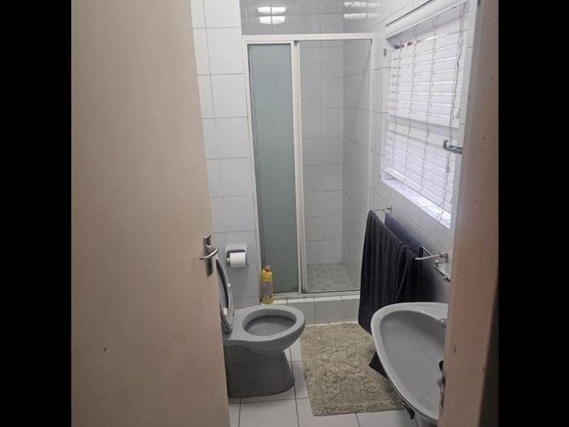A compact bathroom featuring a shower enclosure, pedestal basin, and toilet with white wall tiling and a window.
