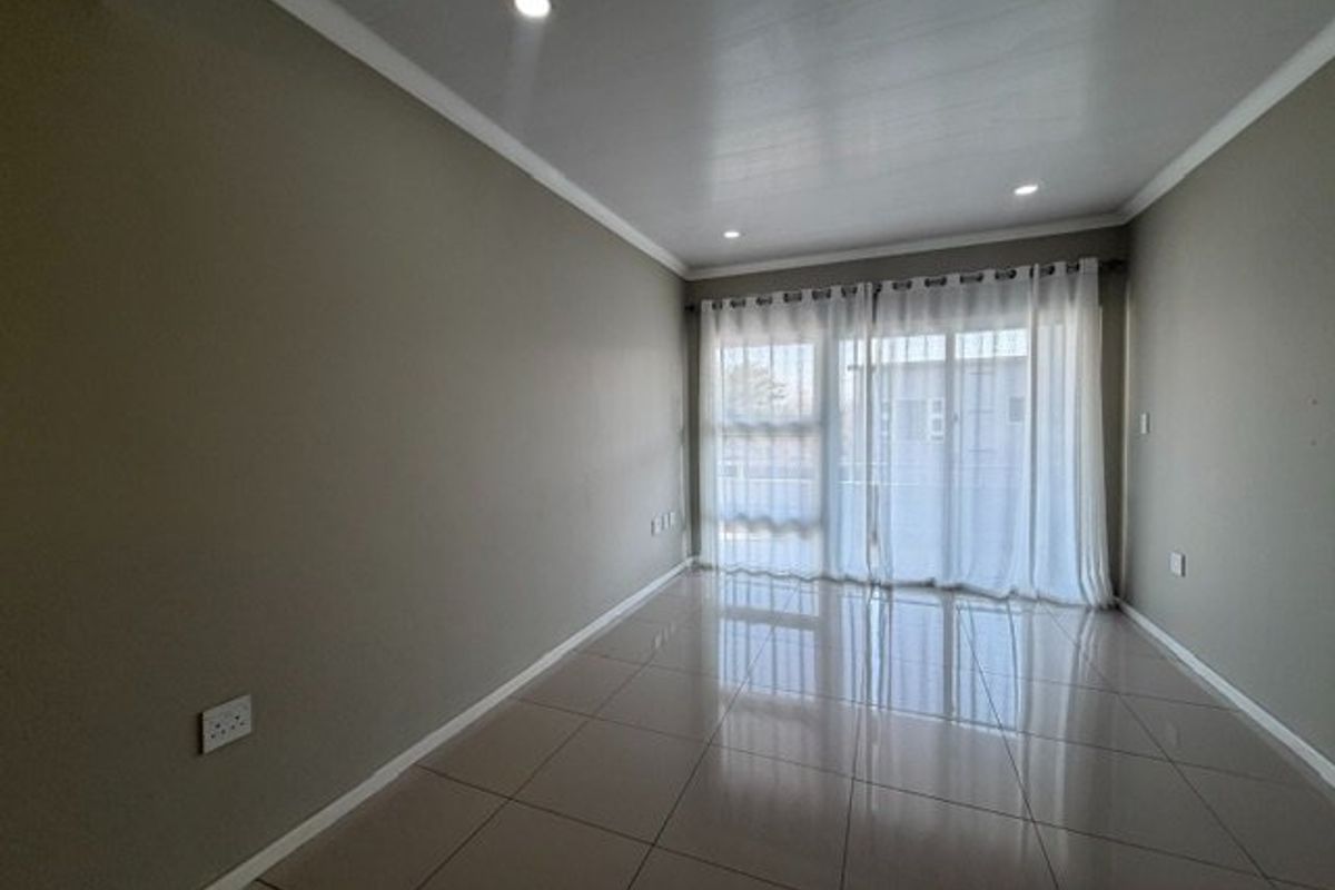 A bright, empty living room featuring neutral-toned tiled flooring, recessed downlighting, and a large sliding glass door with sheer curtains.