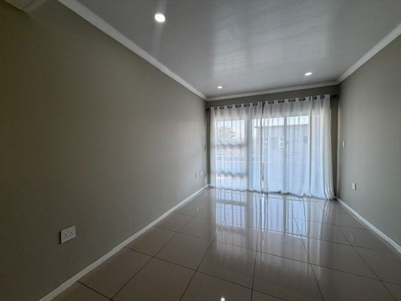 A bright, empty living room featuring neutral-toned tiled flooring, recessed downlighting, and a large sliding glass door with sheer curtains.