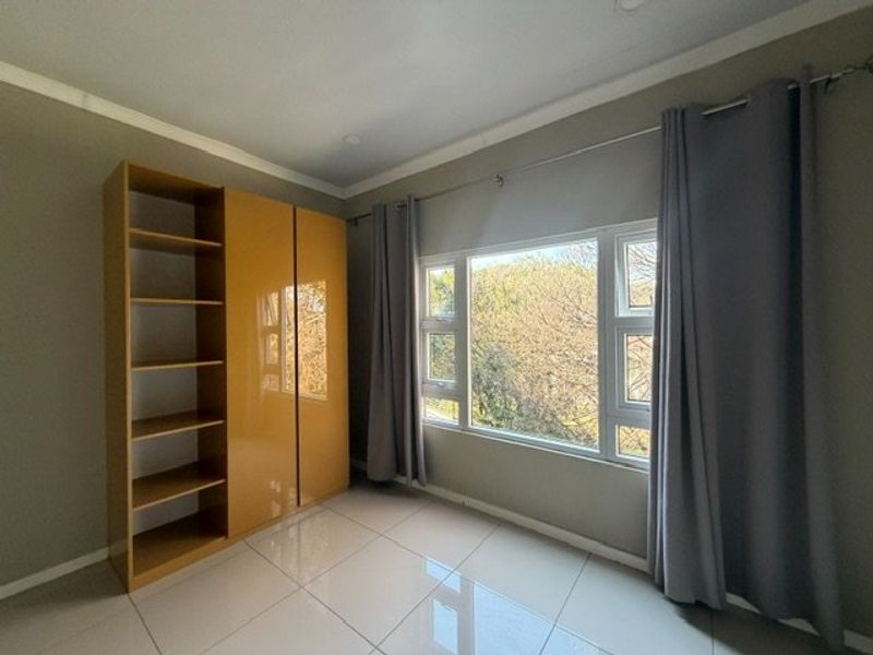 A bright, empty bedroom featuring tiled flooring, a built-in wardrobe with shelving, and large windows with curtain rails.