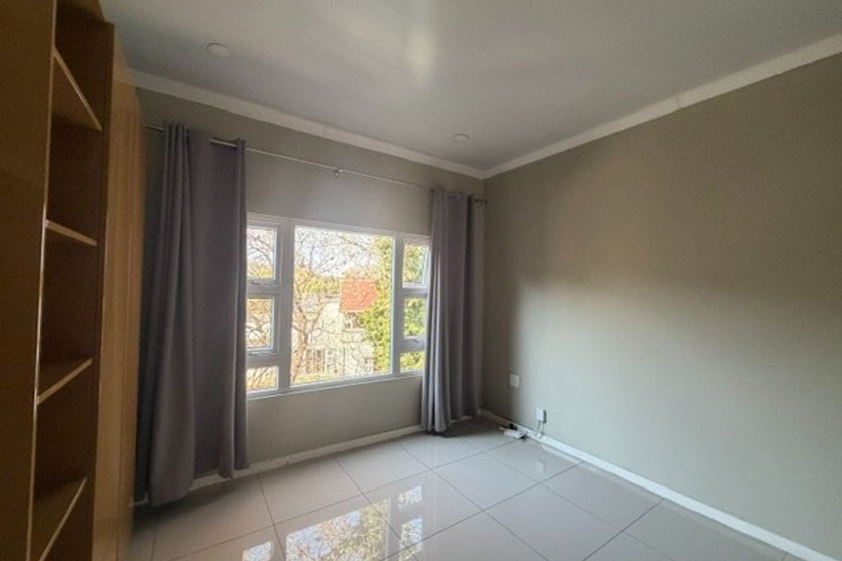 A bright, empty bedroom featuring neutral tiled flooring, built-in cupboards, and large aluminium-framed windows allowing for natural light.