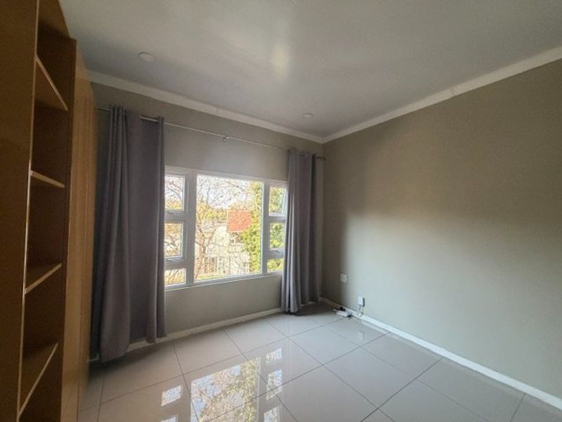 A bright, empty bedroom featuring neutral tiled flooring, built-in cupboards, and large aluminium-framed windows allowing for natural light.