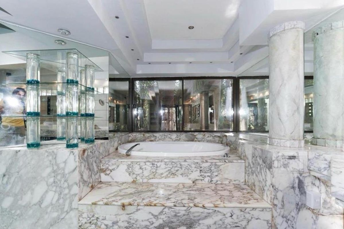 A spacious bathroom featuring a sunken bathtub surrounded by extensive marble cladding, a decorative marble column, and large mirrored wall panels.
