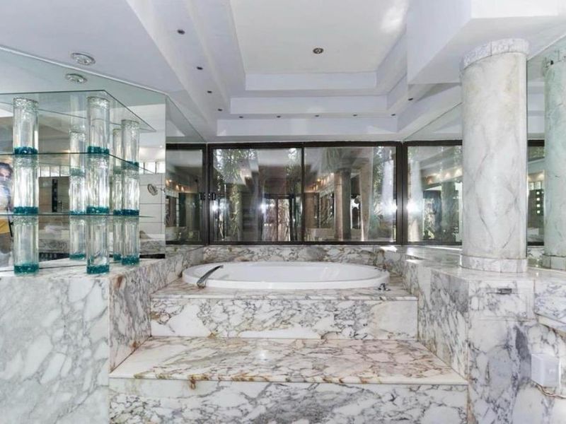 A spacious bathroom featuring a sunken bathtub surrounded by extensive marble cladding, a decorative marble column, and large mirrored wall panels.