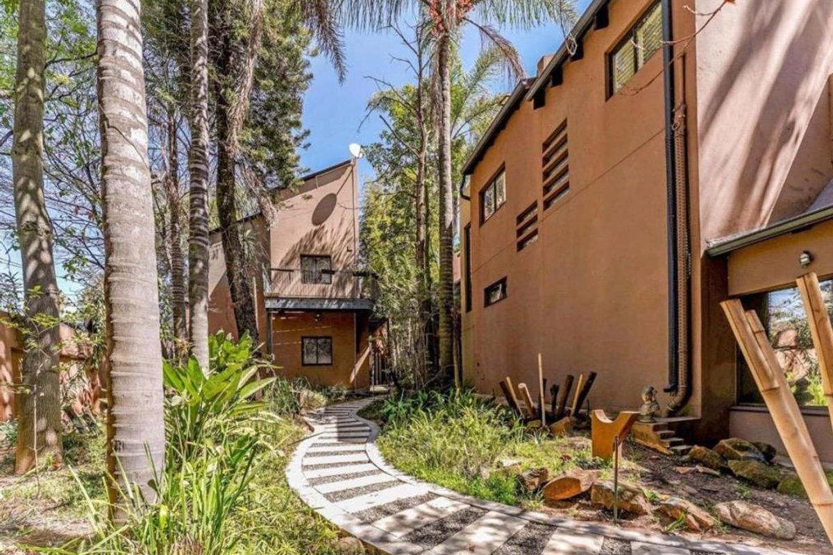 A lush, landscaped rear garden featuring a winding paved pathway leading between two multi-story residential structures with balconies and mature trees.