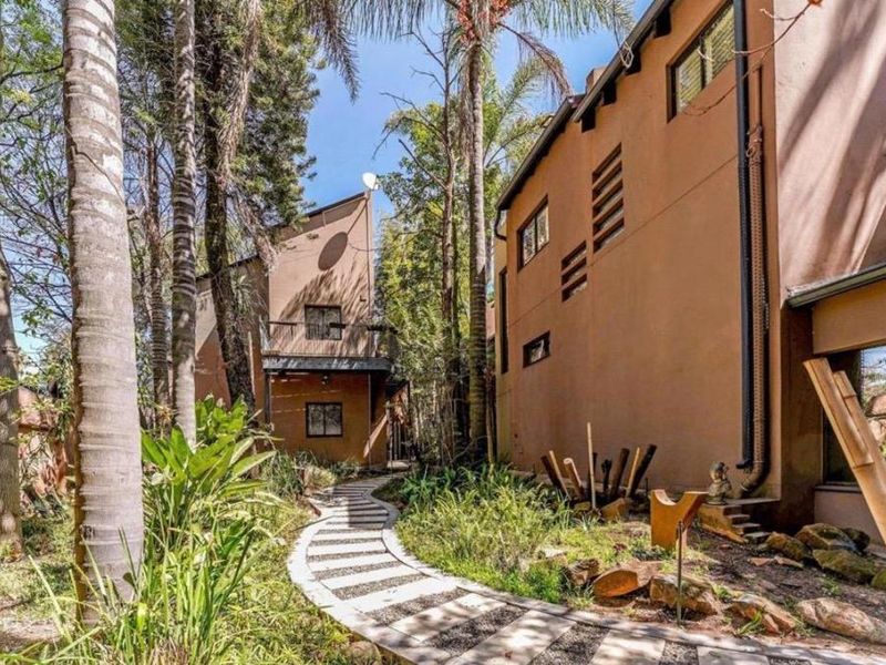 A lush, landscaped rear garden featuring a winding paved pathway leading between two multi-story residential structures with balconies and mature trees.