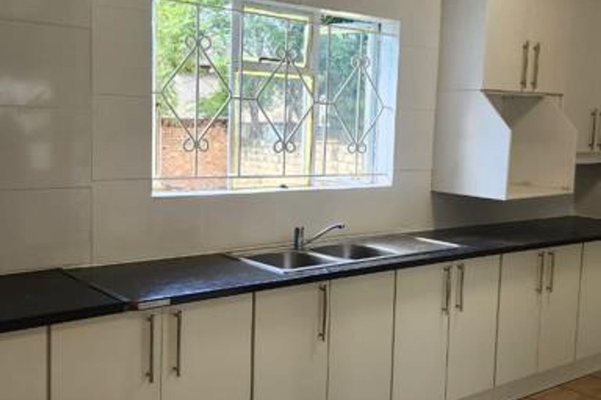 A functional kitchen featuring white built-in cabinetry, dark laminate countertops, a stainless steel double sink, and tiled flooring with security-barred windows.