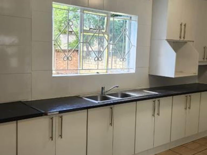 A functional kitchen featuring white built-in cabinetry, dark laminate countertops, a stainless steel double sink, and tiled flooring with security-barred windows.