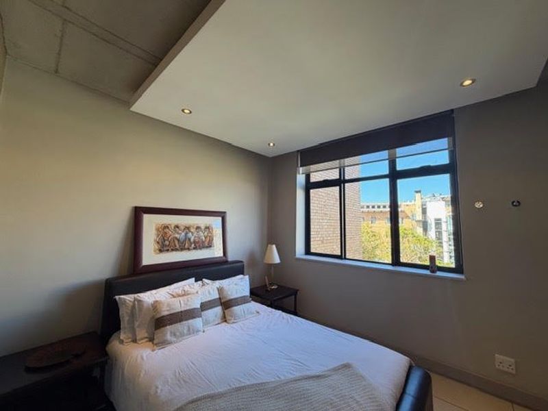 A well-lit bedroom featuring a modern drop ceiling with recessed lighting, large windows with roller blinds, and neutral wall finishes.